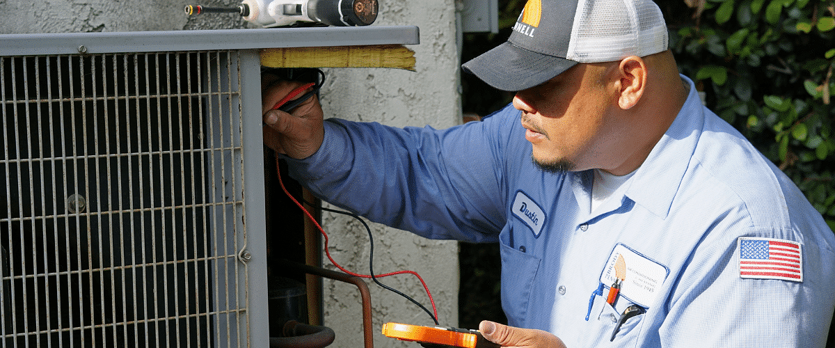 a HVAC pro from Brody Pennel in a light blue service uniform is using a manifold gauge to perform an AC repair on a unit